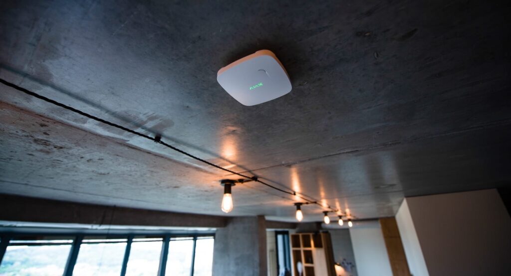 A white Ajax FireProtect wireless fire detector mounted on a raw concrete ceiling in a modern industrial-style apartment, situated next to stylish track lighting with Edison bulbs.