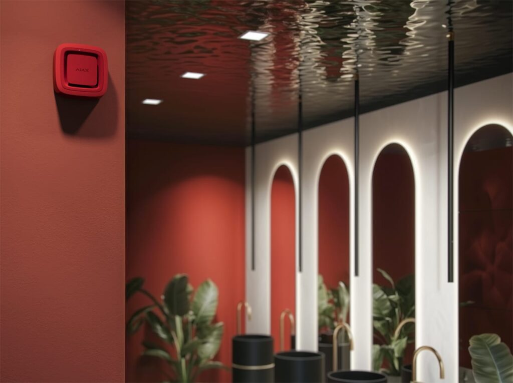 A customized red Ajax smart security device mounted on a deep red wall in a luxurious bathroom setting with a water-ripple effect ceiling and gold fixtures, demonstrating high-end interior design integration.