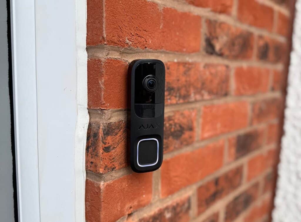 A black Ajax smart video doorbell securely mounted on a red brick wall next to a white door frame, showcasing the camera lens and LED button.
