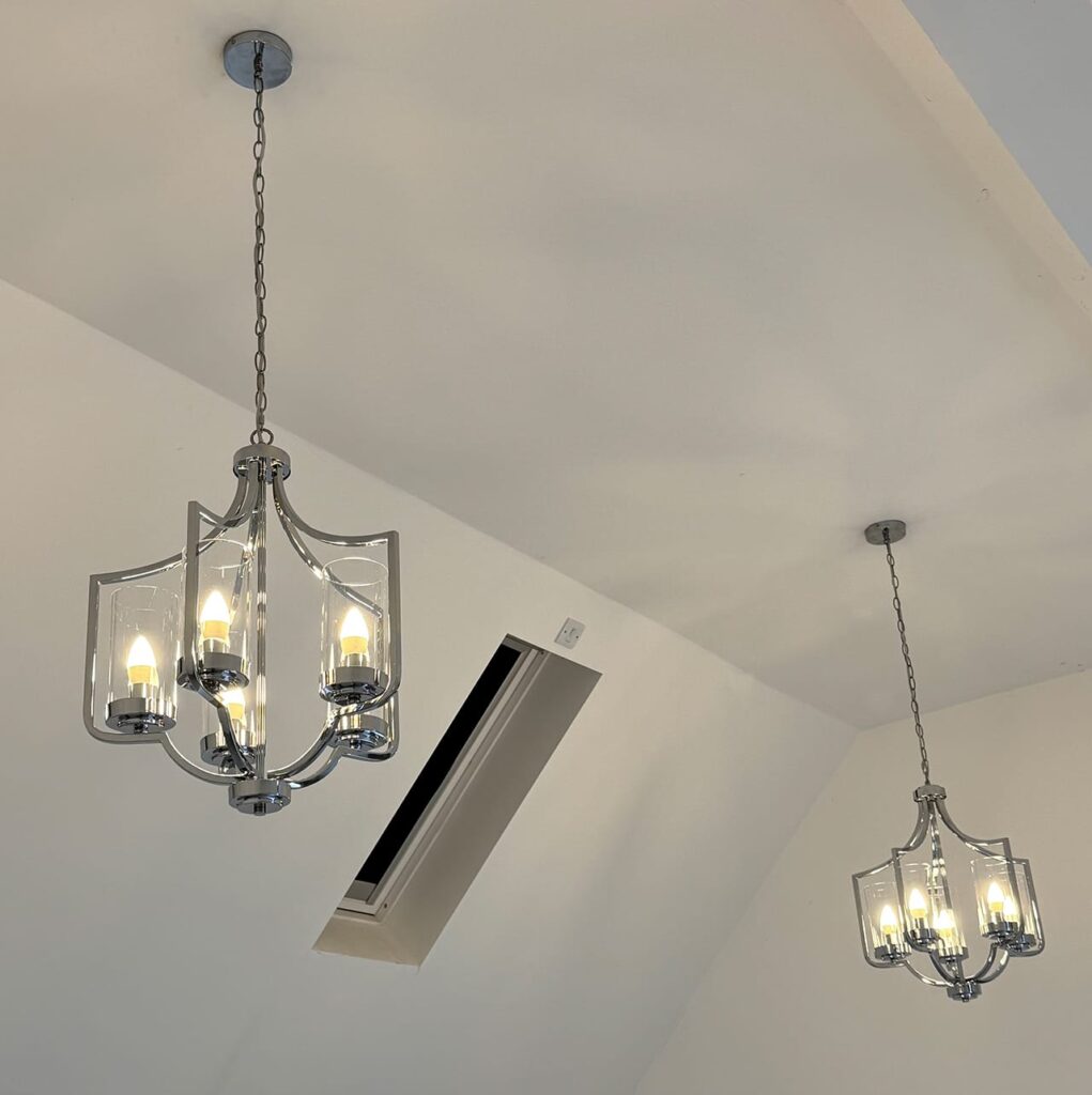 Two modern chrome and glass pendant chandeliers installed on a white sloped ceiling featuring a skylight, showcasing high-end residential electrical lighting design.