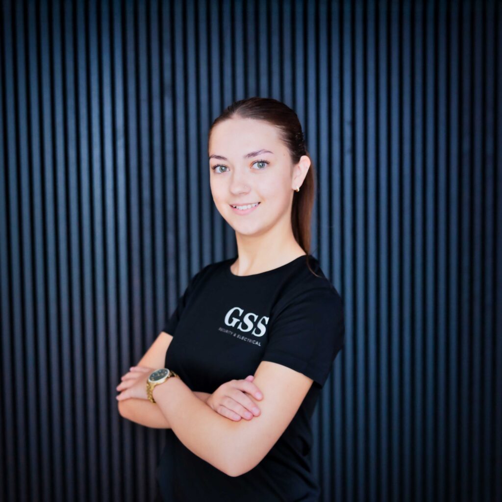 A professional headshot of a GSS Security & Electrical team member named Amelia, wearing a black branded t-shirt and smiling with arms crossed against a modern dark vertical slat background.