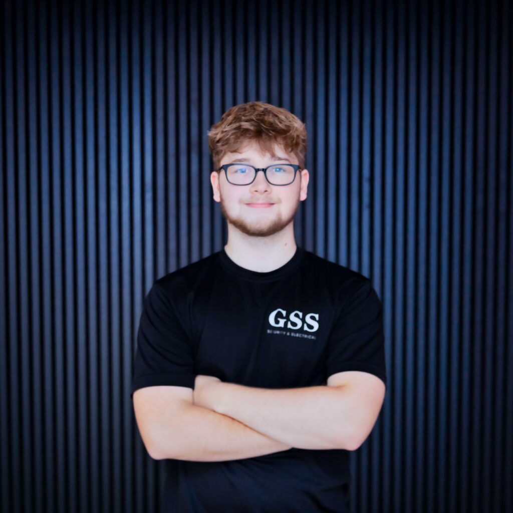 A professional headshot of a GSS Security & Electrical team member named Bayley, wearing glasses and a black branded t-shirt, smiling with arms crossed against a dark vertical slat background.