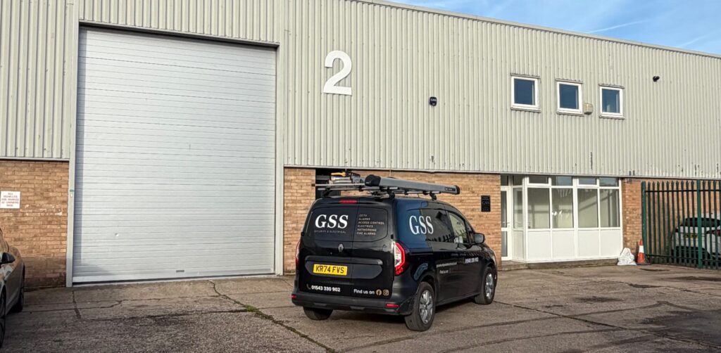 A black GSS Security & Electrical van parked outside the Just Blinds showroom in a sunny commercial business park.