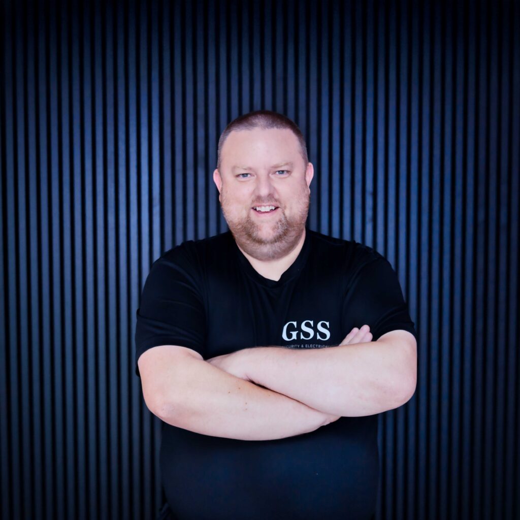 A professional headshot of a GSS Security & Electrical team member named Dave, smiling with arms crossed and wearing a black branded t-shirt against a dark vertical slat background.