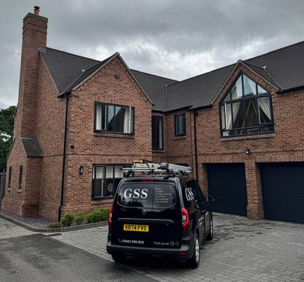 A black GSS Security & Electrical van parked on the paved driveway of a large modern detached house, equipped with roof ladders for a residential security installation project.