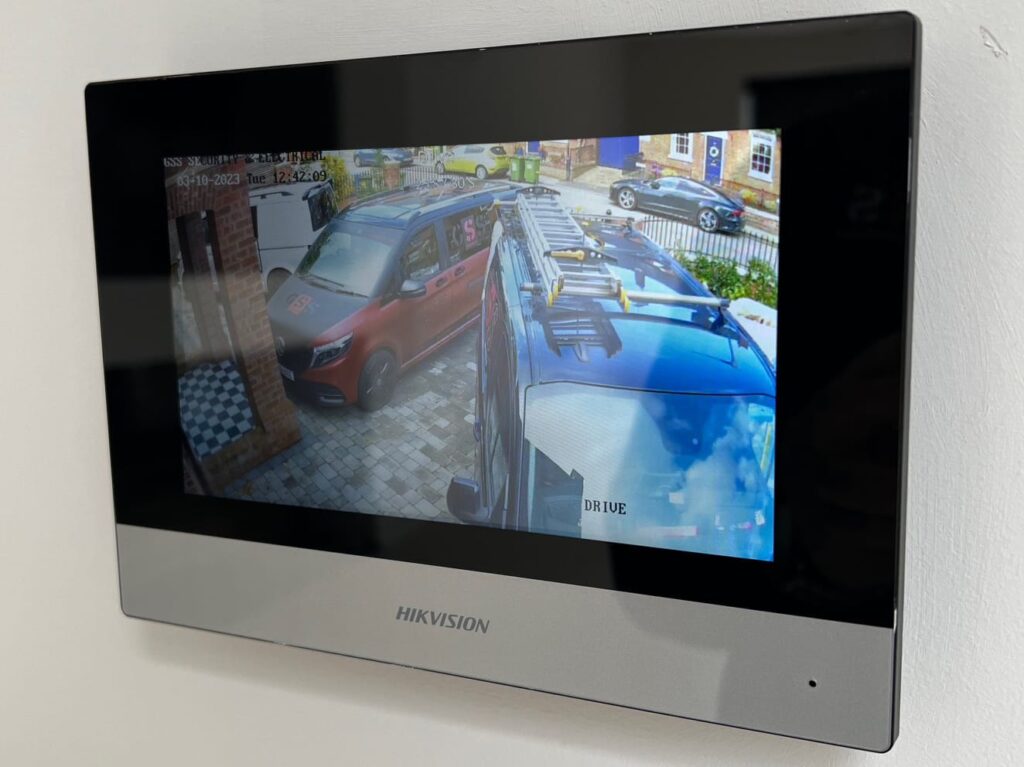 A wall-mounted Hikvision indoor security monitor displaying live high-definition CCTV footage of company vans parked on a residential driveway.
