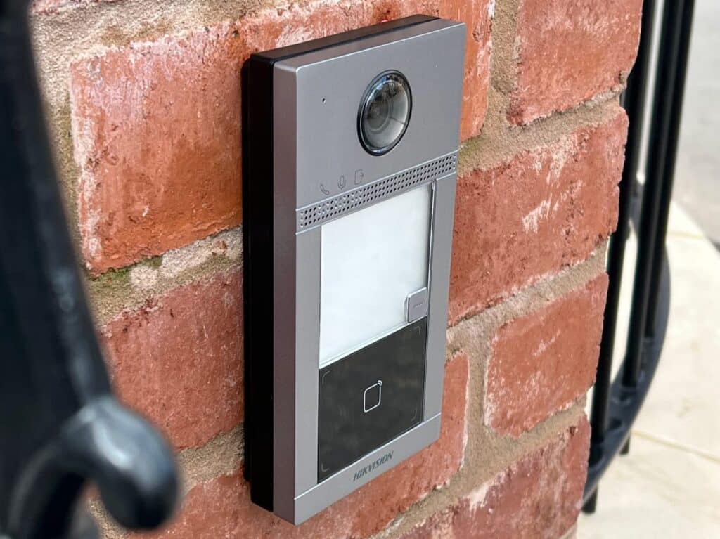 A silver Hikvision video intercom door station professionally mounted on a red brick wall, featuring a built-in camera, speaker, and card reader for secure access control.
