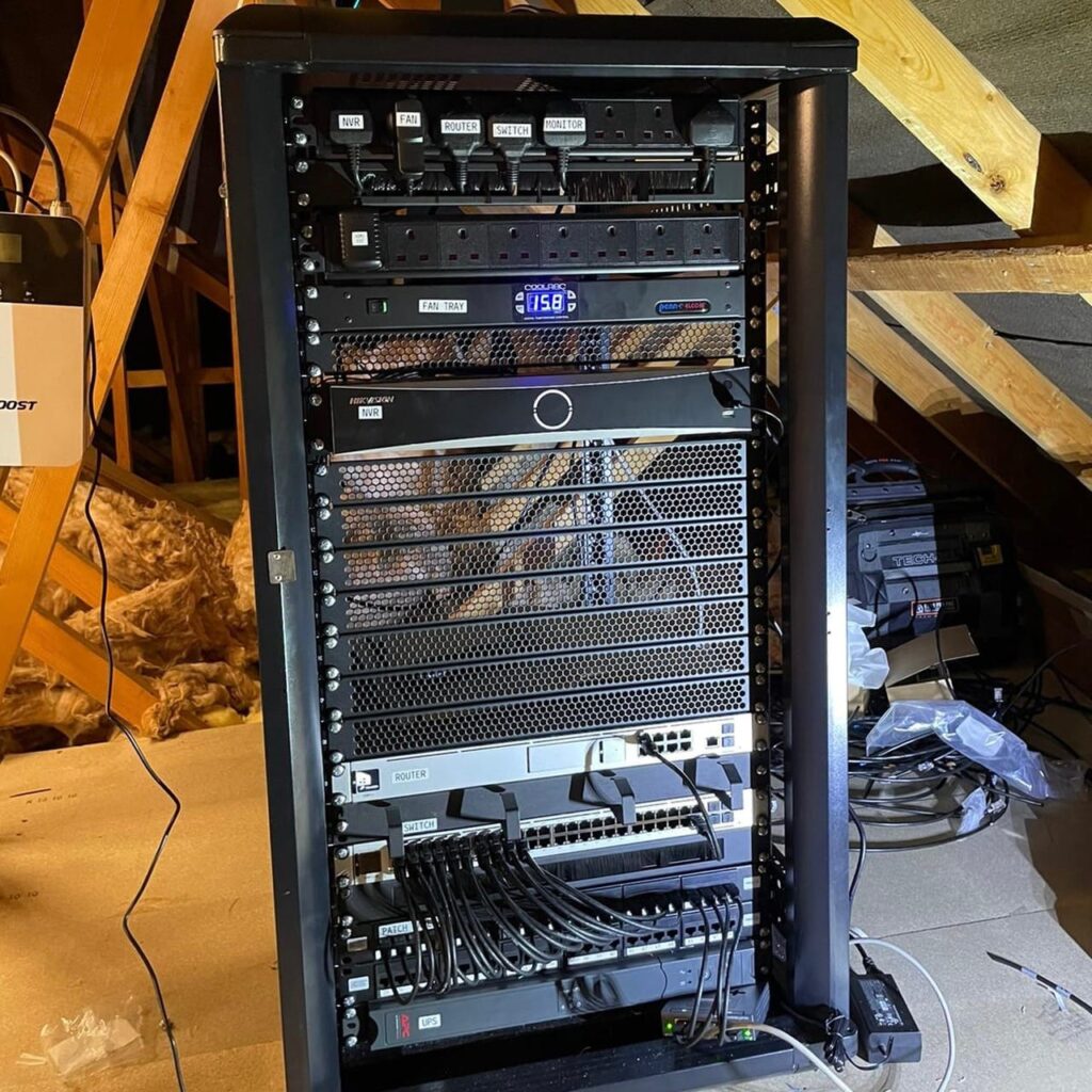 A professionally installed black data cabinet located in a residential loft, housing a Hikvision NVR, network switches, patch panels, and a cooling tray with neat cable management for smart home connectivity.
