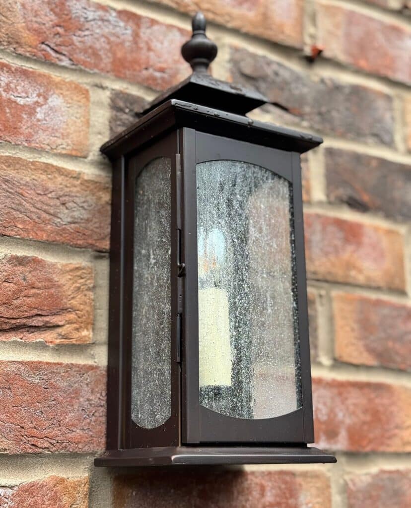 A traditional black outdoor wall lantern featuring seeded glass panels and a candle-style bulb, professionally mounted on a red brick wall to enhance exterior lighting.